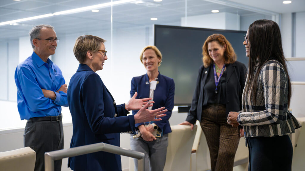 Five professionally dressed people stand in a modern office space, engaged in a friendly conversation. One woman gestures with her hands while speaking, as the others smile and listen.