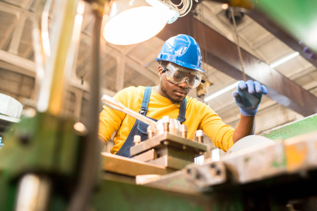 Busy young factory engineer in hardhat and safety goggles examining milling lathe and repairing it while working at production plant
